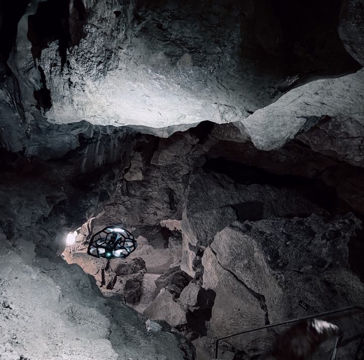 Eine hell erleuchtete Inspektionsdrohne mit Schutzrahmen fliegt durch eine enge, dunkle Felsenhöhle.